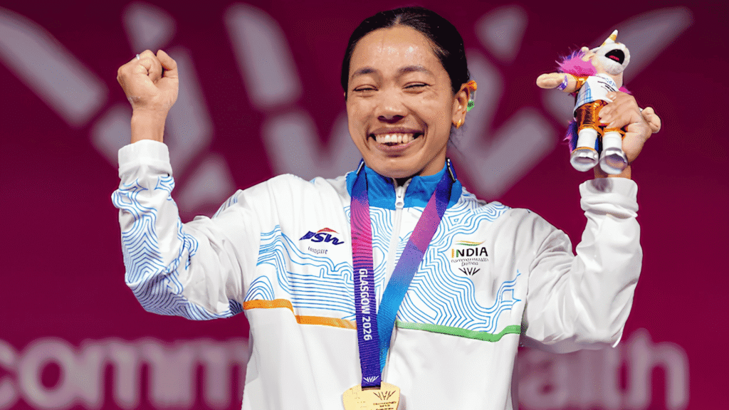 Mirabai Chanu celebrates after winning India's first gold medal at the Commonwealth Games 2026, wearing her gold medal and Team India jacket while raising her fist and holding the Glasgow 2026 mascot.