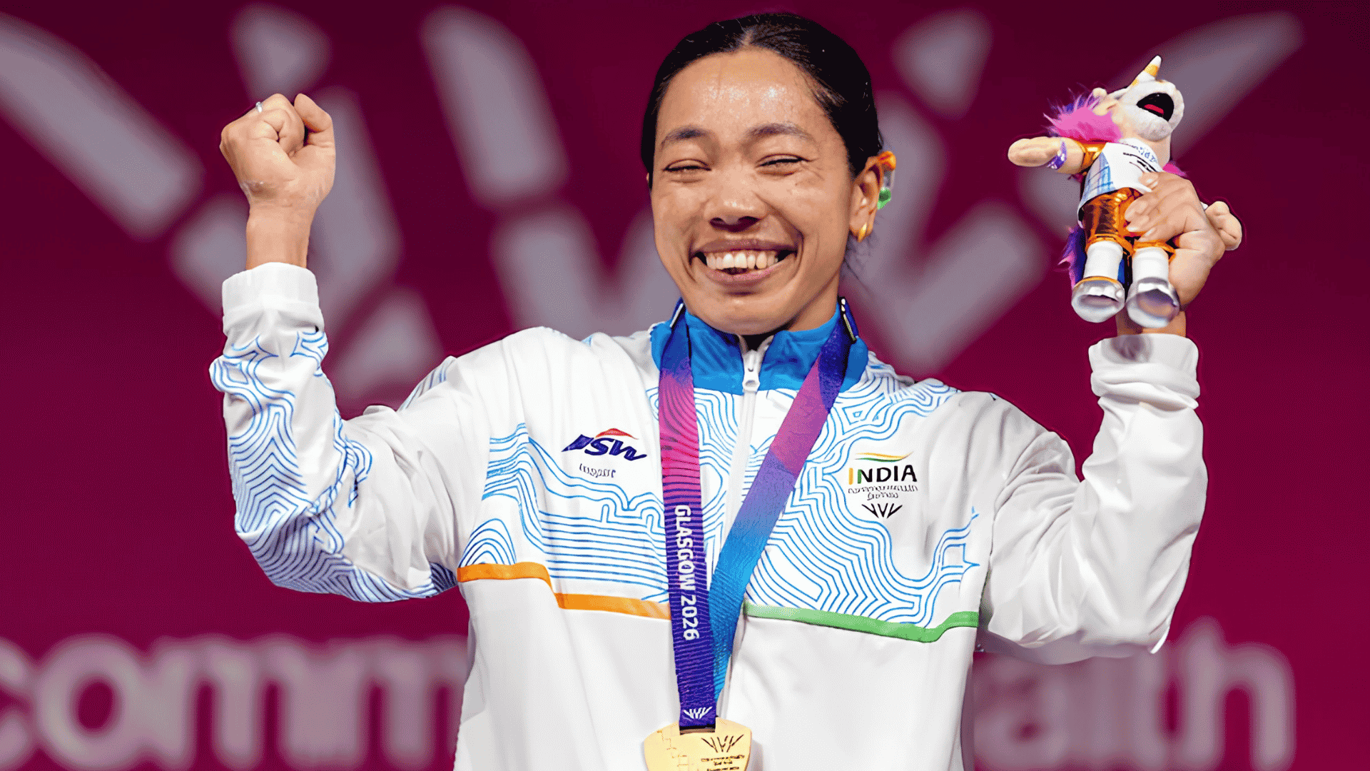 Mirabai Chanu celebrates after winning India's first gold medal at the Commonwealth Games 2026, wearing her gold medal and Team India jacket while raising her fist and holding the Glasgow 2026 mascot.